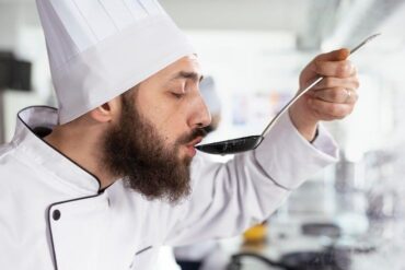 Kock smakar av en sås ur en sked i ett ljust kök